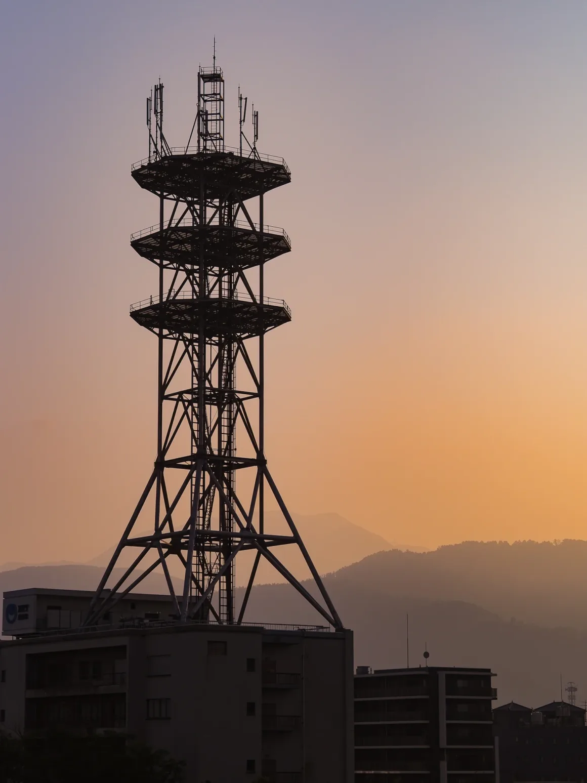 OM System PEN İncelemesi: Tanıdık Ama Son Derece Şık 40 A tall metal telecommunications tower stands silhouetted against a sunset sky, rising above a building with mountains in the distance.