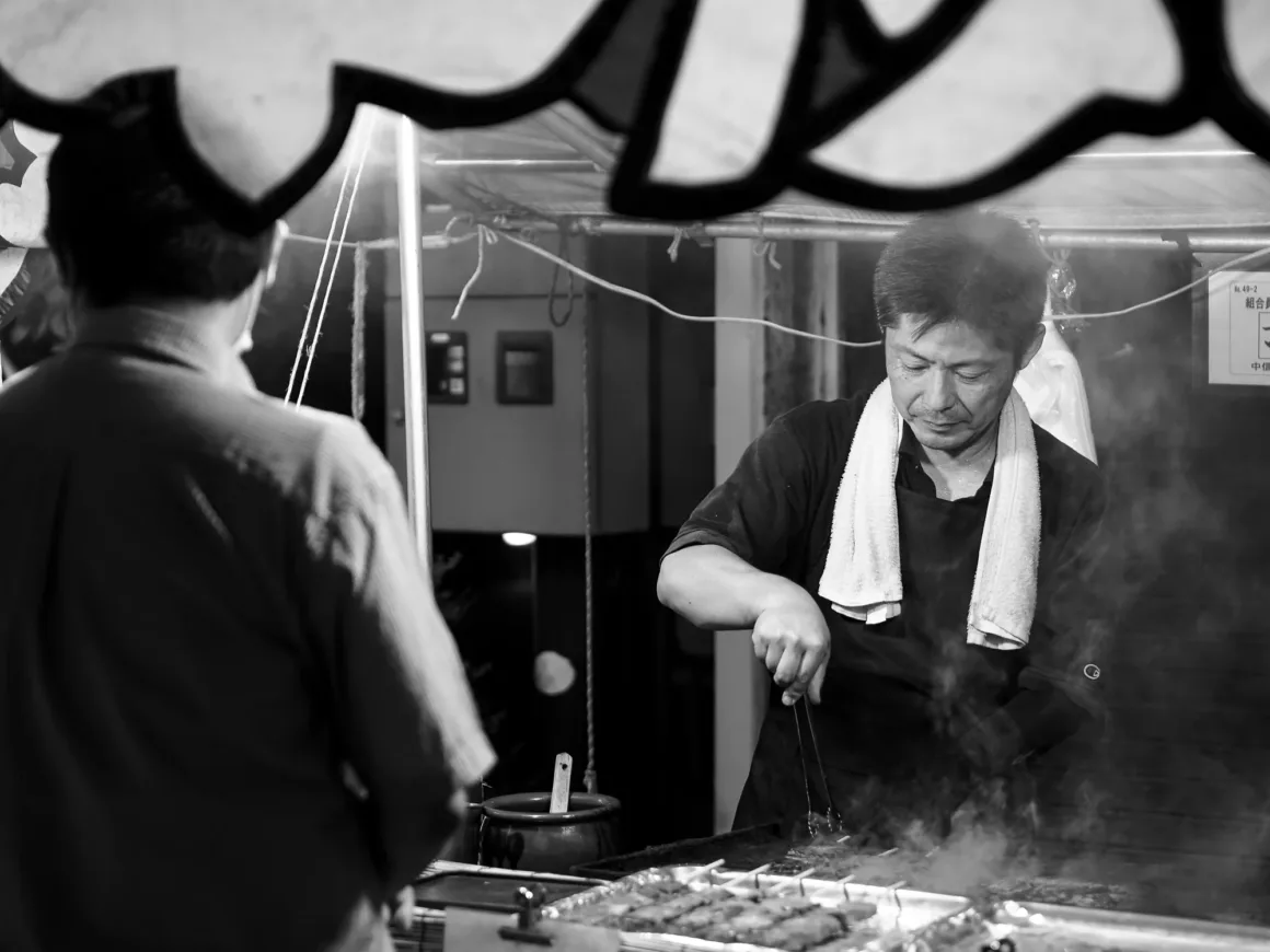OM System PEN İncelemesi: Tanıdık Ama Son Derece Şık 31 A man with a towel around his neck uses tongs to grill skewers at a street food stall while a customer stands in the foreground.