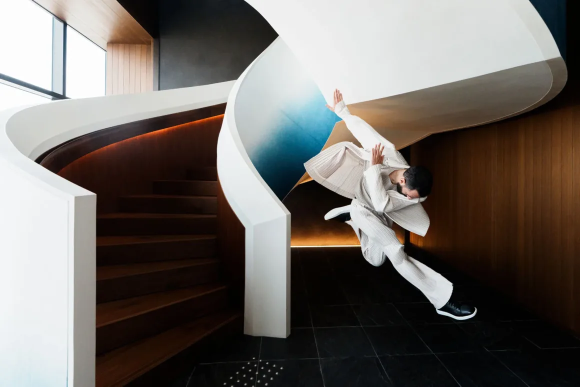 A person in a white pleated outfit leaps in mid-air next to a modern, curved wooden staircase with white railings.