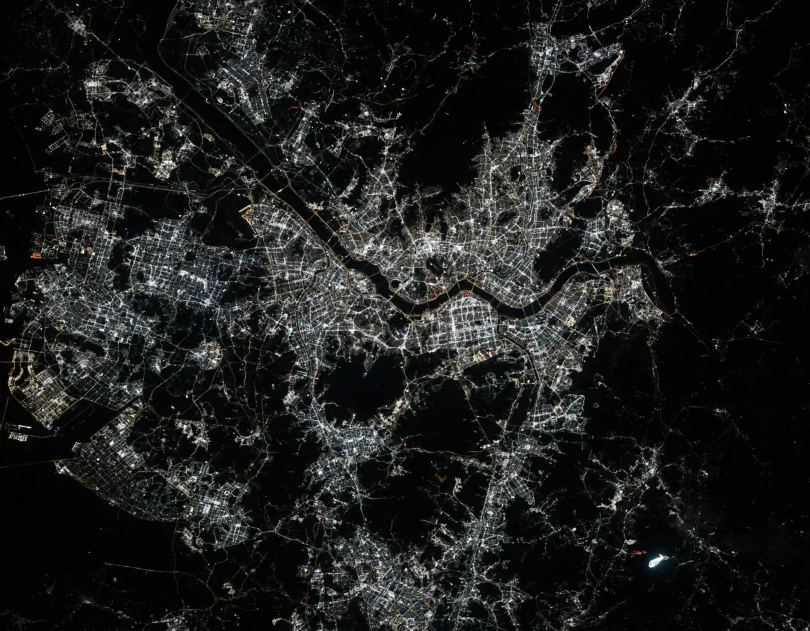 Astronotun Önce-Sonra Fotoğrafları Kentlerin Turuncudan Beyaz Işığa Geçişini Gösteriyor 26 A satellite view of a brightly lit city at night, featuring a prominent river winding through the dense urban grid.