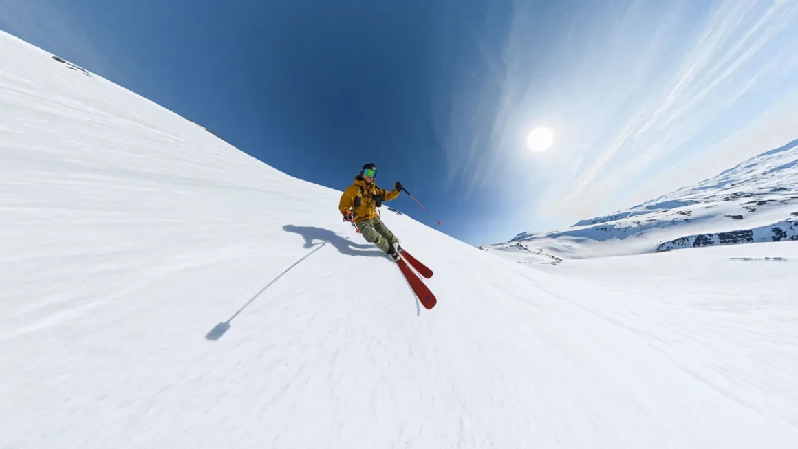 A skier in a yellow jacket and green pants carves down a bright, snow-covered mountain slope under a clear blue sky.