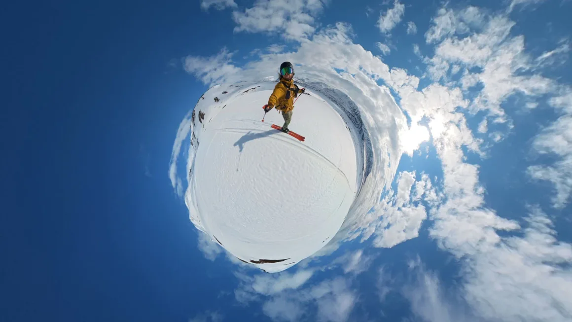 A skier in a yellow jacket and helmet stands on a snowy slope in a 360-degree panoramic view under a bright blue sky with clouds.