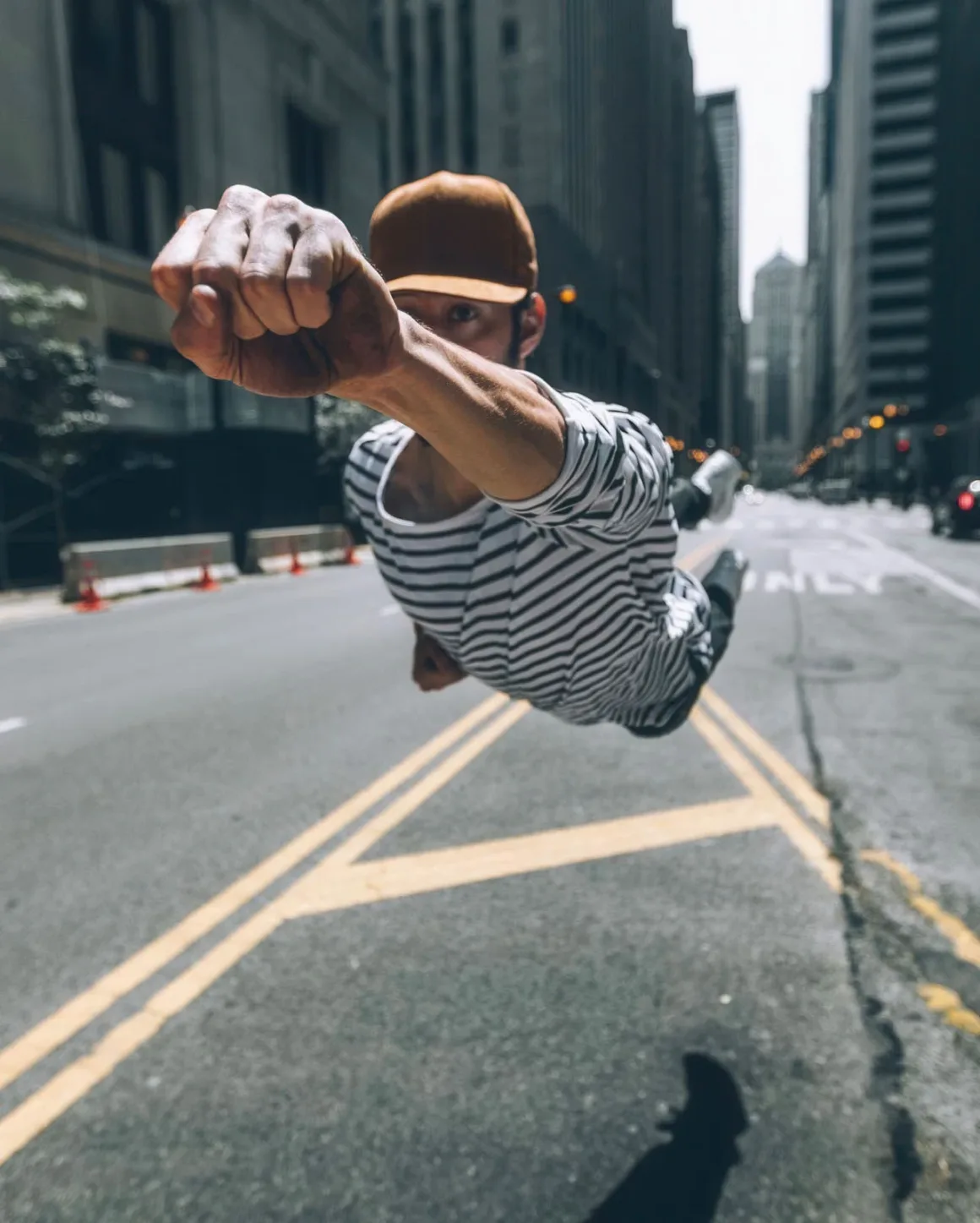 A person wearing a striped shirt and a brown cap appears to be flying horizontally above a city street, with their fist extended forward.