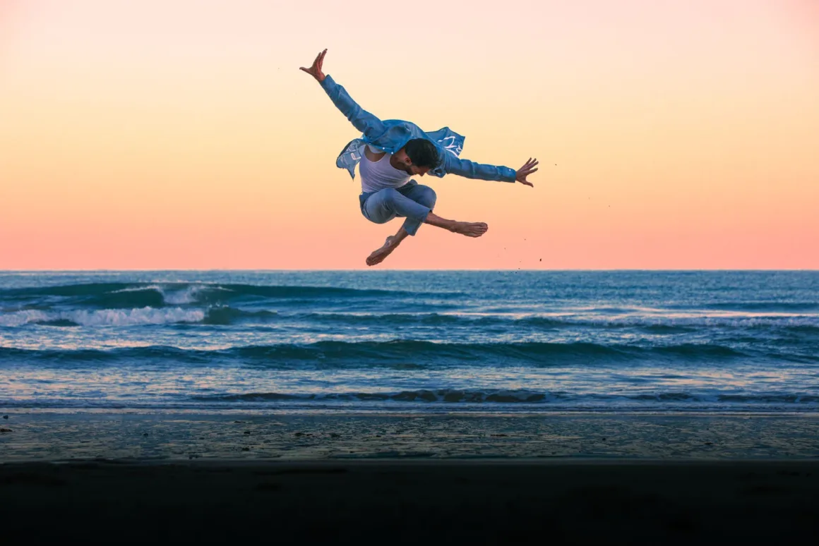 A man in a white tank top and blue shirt leaps mid-air against a sunset sky above the ocean.