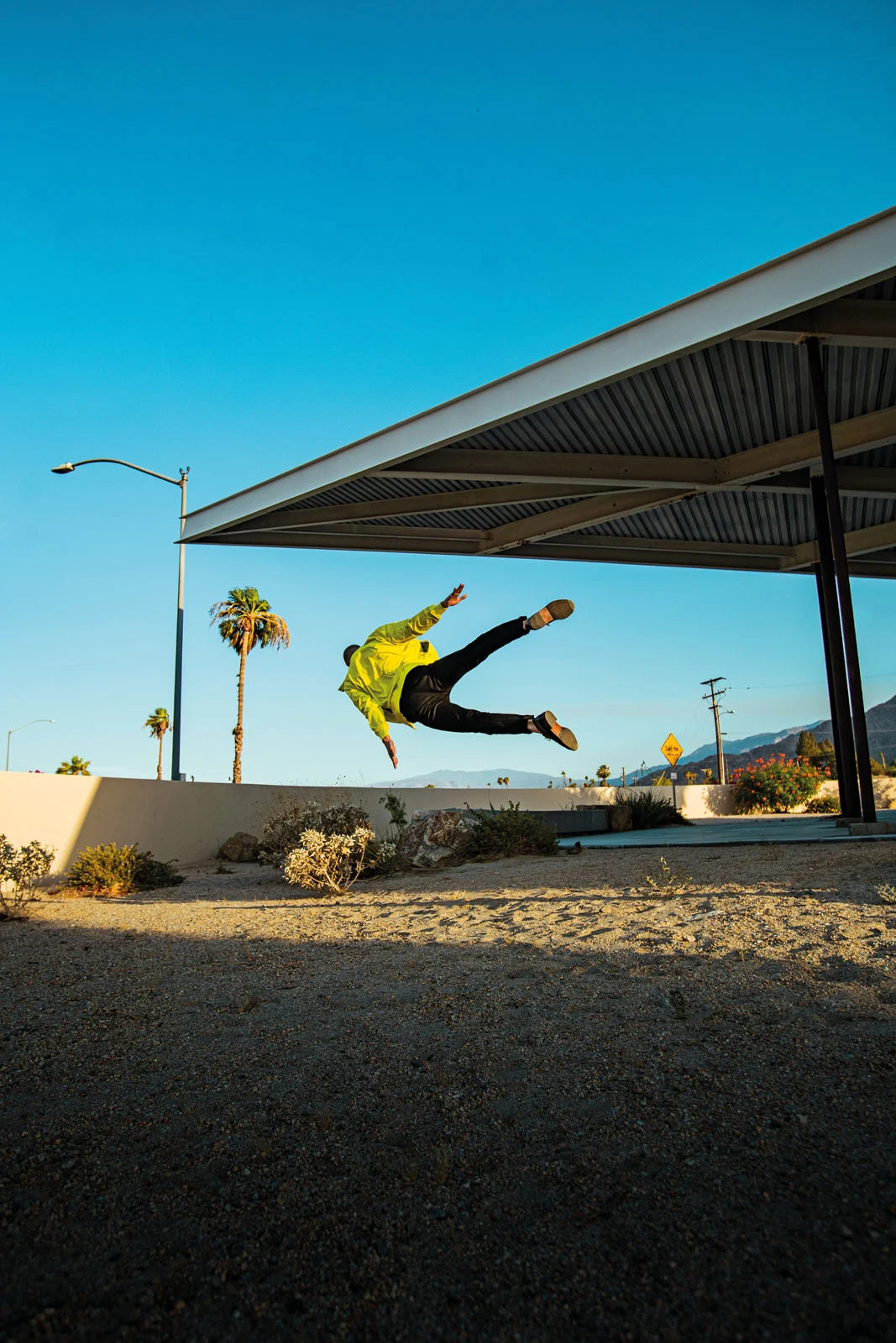 A person in a bright yellow jacket and black pants leaps horizontally through the air beneath a modern architectural overhang.