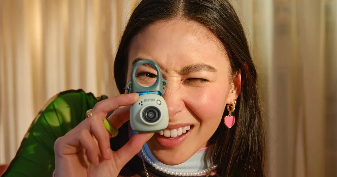 Fujifilm Instax Pal 2, Tuhaf Selefinden Tamamen Farklı Görünüyor 19 Uzun koyu saçlı bir kişi, açık yeşil renkli küçük dijital kamerayı gözüne tutarken göz kırpıyor.