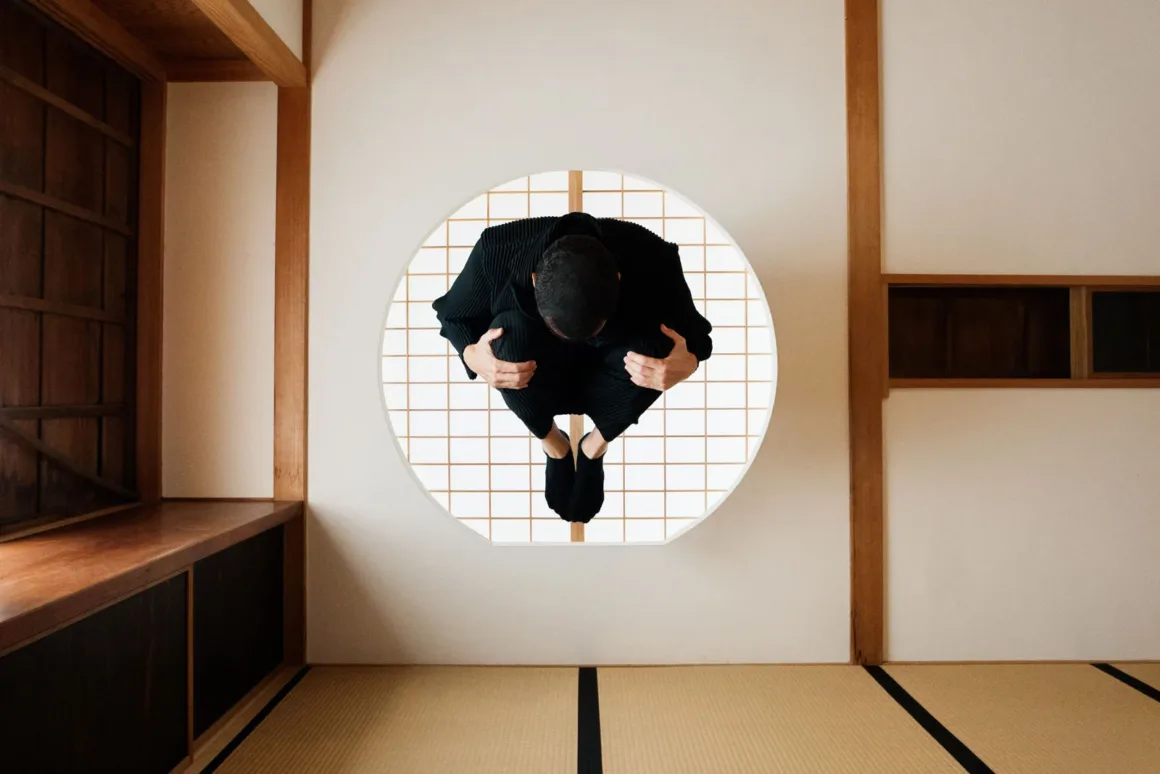 A person in black clothing is crouched in a fetal position, appearing to float in front of a circular window with a grid.