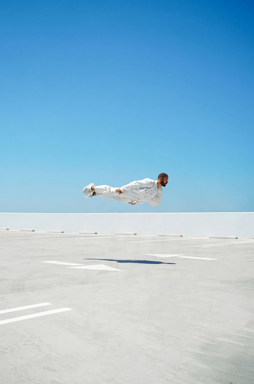 A man wearing all-white clothing floats horizontally in mid-air above a parking lot under a clear blue sky.