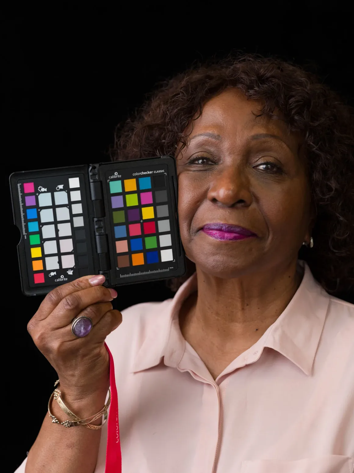 Fotoğrafçıların Renkli Dostu ColorChecker 50 Yaşında 22 Kıvırcık koyu saçlı ve açık pembe gömlekli bir kadın, ColorChecker Classic kartını yüzünün yanında tutuyor.