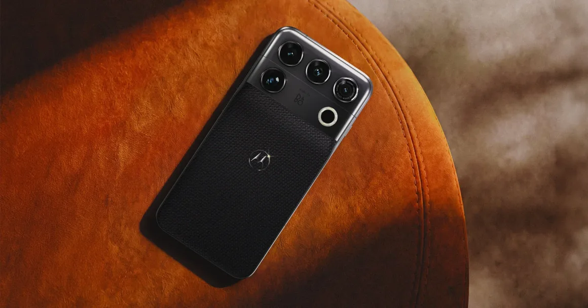 Motorola Signature 27, Mobil Fotoğrafçılığın Sınırlarını Zorluyor 20 Dokulu arka yüzeye sahip siyah bir akıllı telefon, üç lensli belirgin bir kamera modülü ve dairesel flaşıyla turuncu bir yüzeyin üzerinde duruyor.