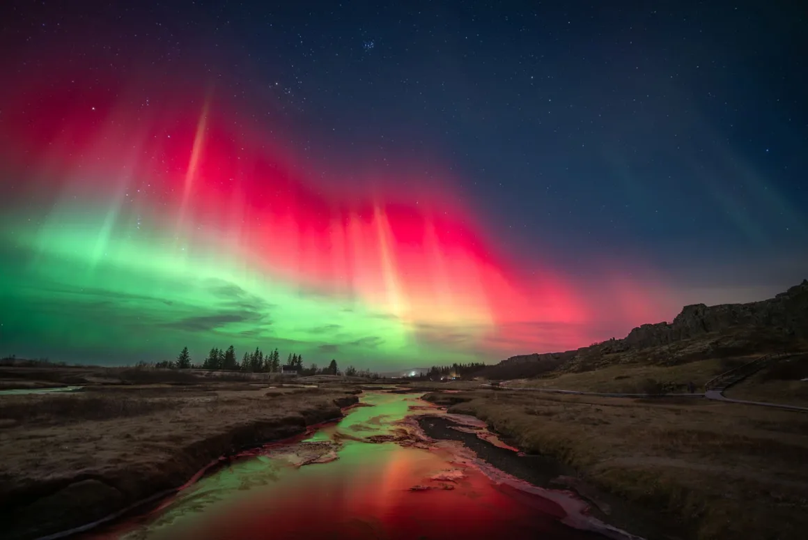 Astronomy Photographer of the Year 2026: Bu Dünyadan Olmayan 31 Kazanan 25 Vibrant red and green aurora borealis lights illuminate the night sky above a calm stream flowing through a grassy landscape.