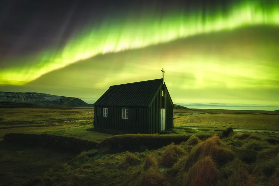 Astronomy Photographer of the Year 2026: Bu Dünyadan Olmayan 31 Kazanan 24 A small, dark wooden church with a cross on its roof stands in a grassy field under a vibrant green aurora borealis.