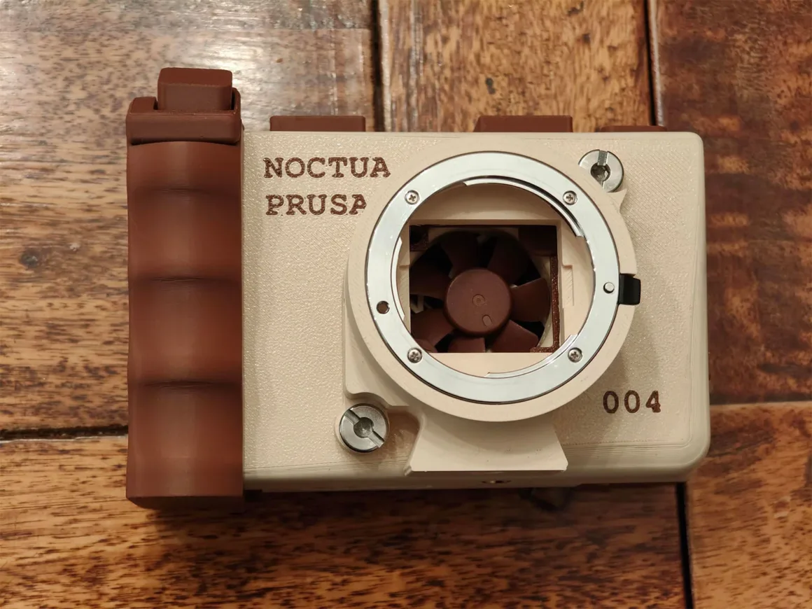 Noctua’nın bej ve kahverengi renklerini taşıyan 3D baskılı fotoğraf makinesi gövdesi; objektif yuvasının içinden küçük bir soğutma fanı görülüyor ve ön yüzünde "NOCTUA PRUSA" ile "004" yazıları yer alıyor.