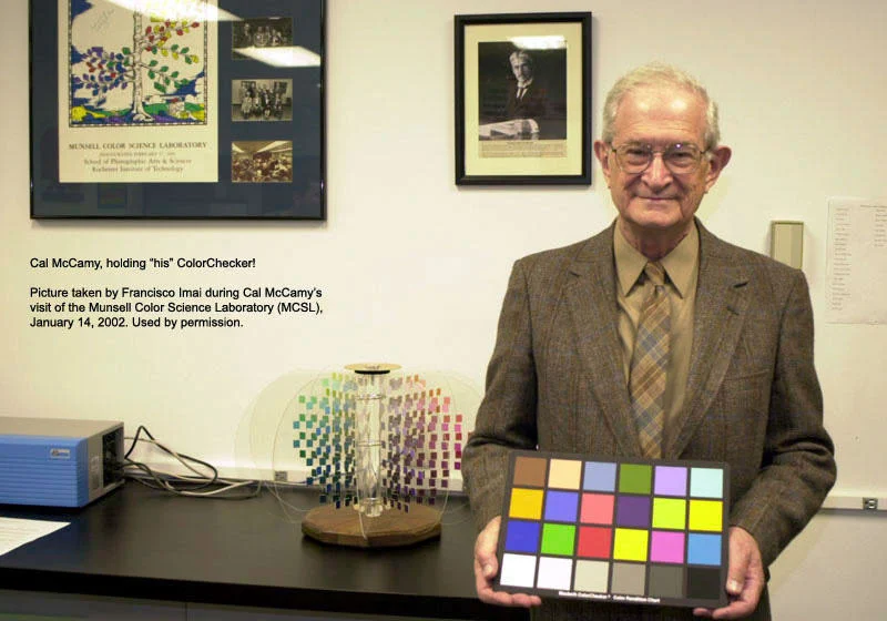 Fotoğrafçıların Renkli Dostu ColorChecker 50 Yaşında 18 Kahverengi takım elbiseli Dr. Cal McCamy, masasının önünde renk kontrol kartını tutuyor.
