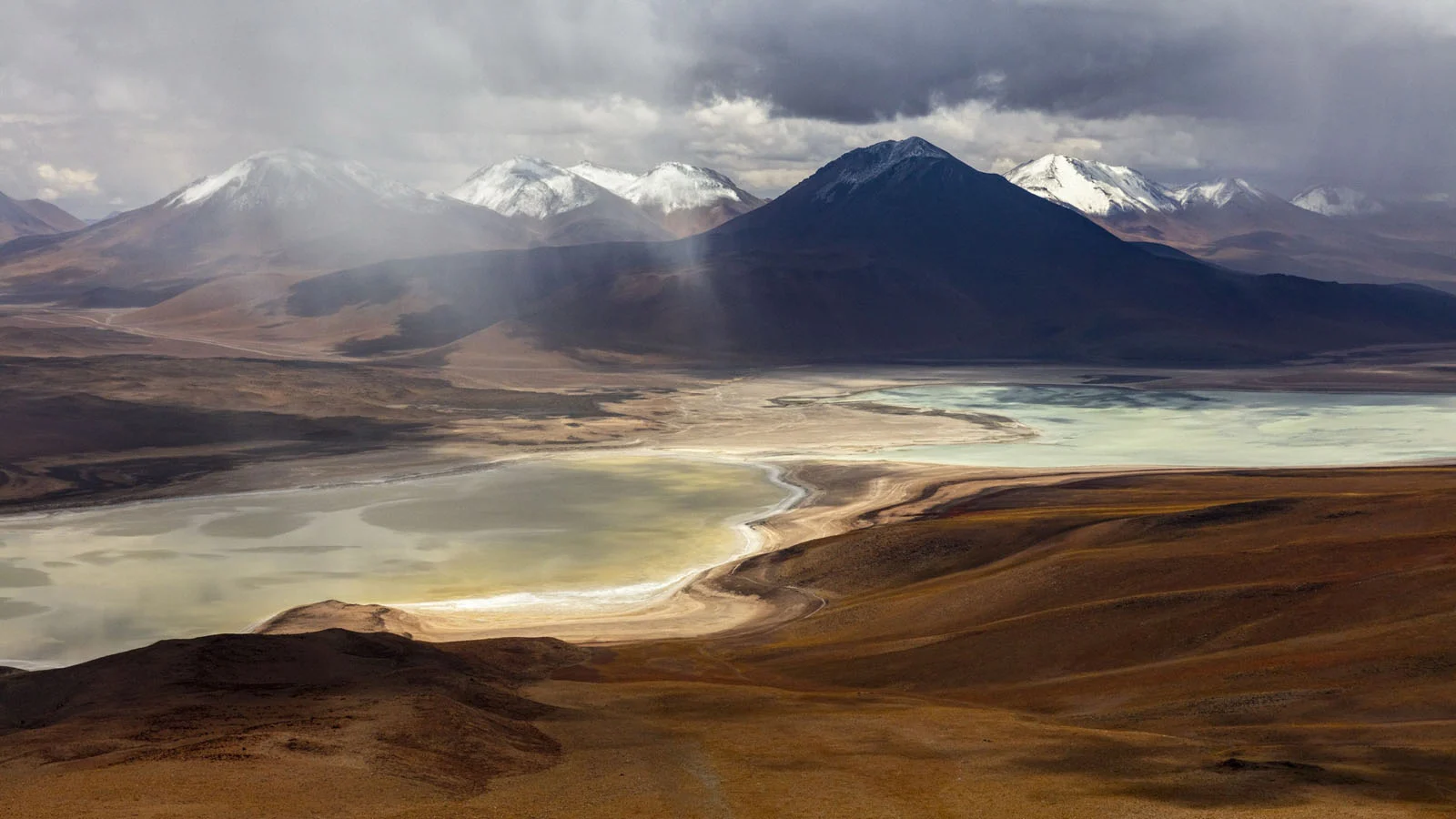 Natural Landscape Photography Awards 2026’nın Kazananları Açıklandı 70 A high-angle view of a pale, shallow lake surrounded by arid, brown hills under a dramatic, cloudy sky with snow-capped mountains.