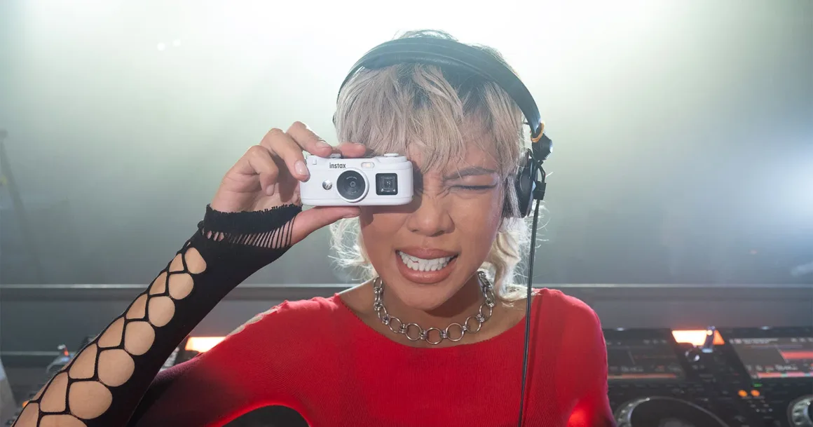 Fujifilm Instax Pal 2, Tuhaf Selefinden Tamamen Farklı Görünüyor 23 A person with blonde hair and headphones holds a small white camera up to their eye while smiling in a DJ booth.