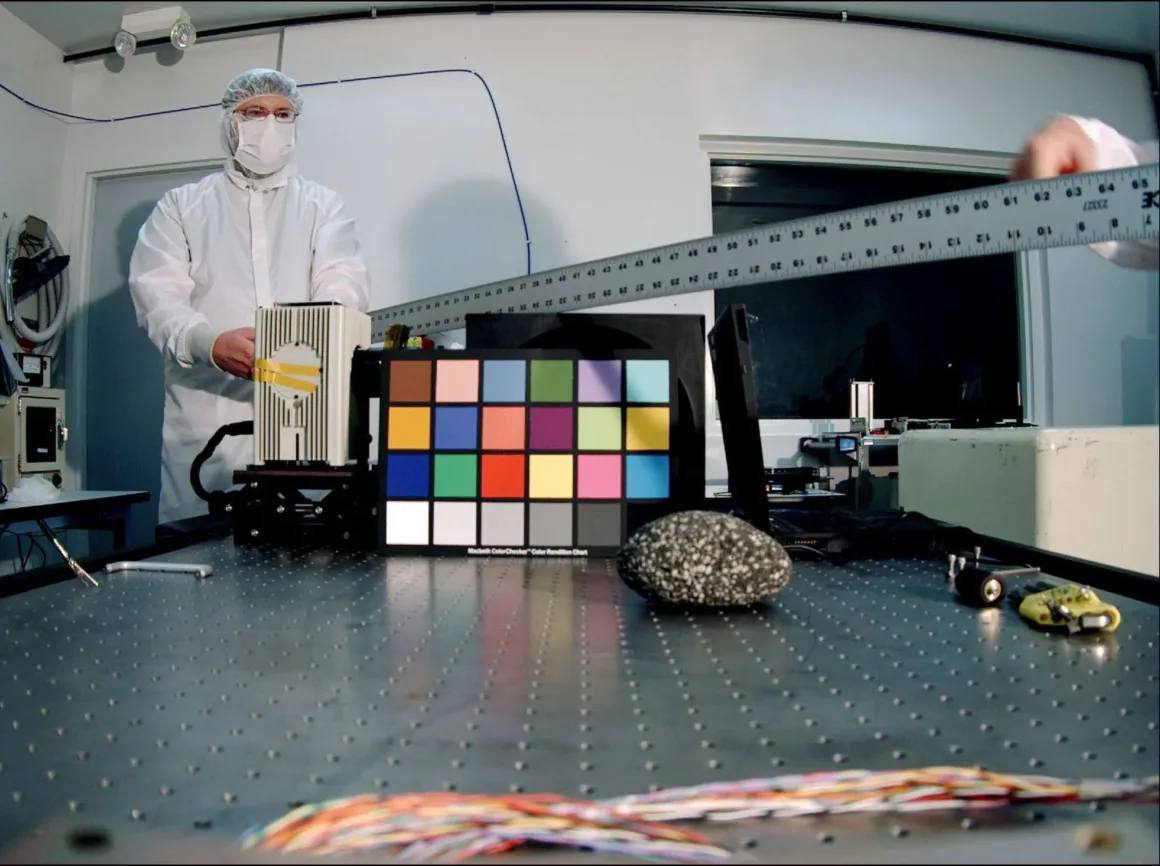 Fotoğrafçıların Renkli Dostu ColorChecker 50 Yaşında 26 Beyaz temiz oda kıyafeti giymiş bir kişi, masadaki metal cetvel, renk kalibrasyon kartı ve kaya örneğinin arkasında duruyor.