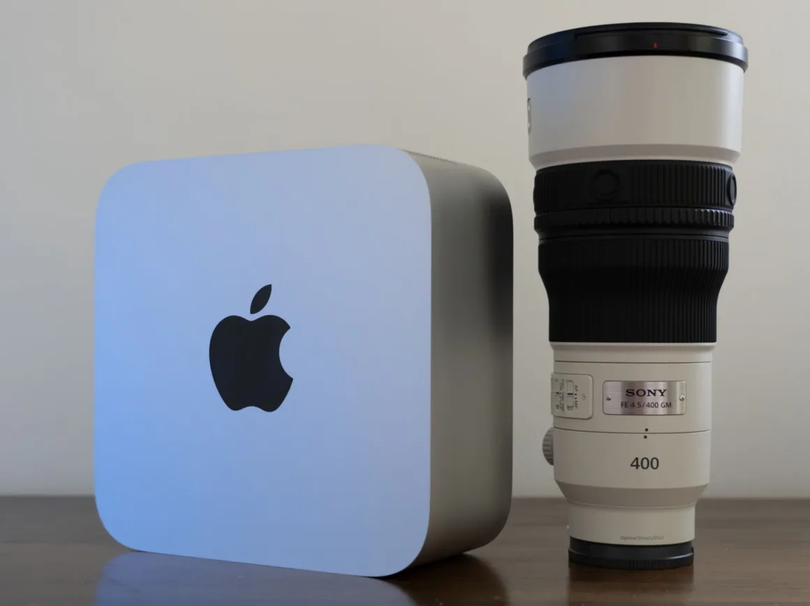 Sade bir duvarın önündeki ahşap yüzeyde Sony FE 400mm F4.5 GM objektifin yanında duran Apple Mac Studio