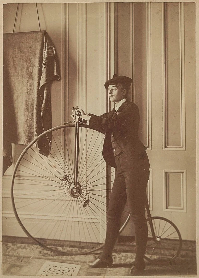 “Tehlikeli” Kadın Fotoğrafçılar Yeni Kitapta Buluştu 17 Takım elbise ve şapka giyen kişi, panelli bir duvarın önünde büyük ön tekerlekli penny-farthing bisikletin yanında duruyor.