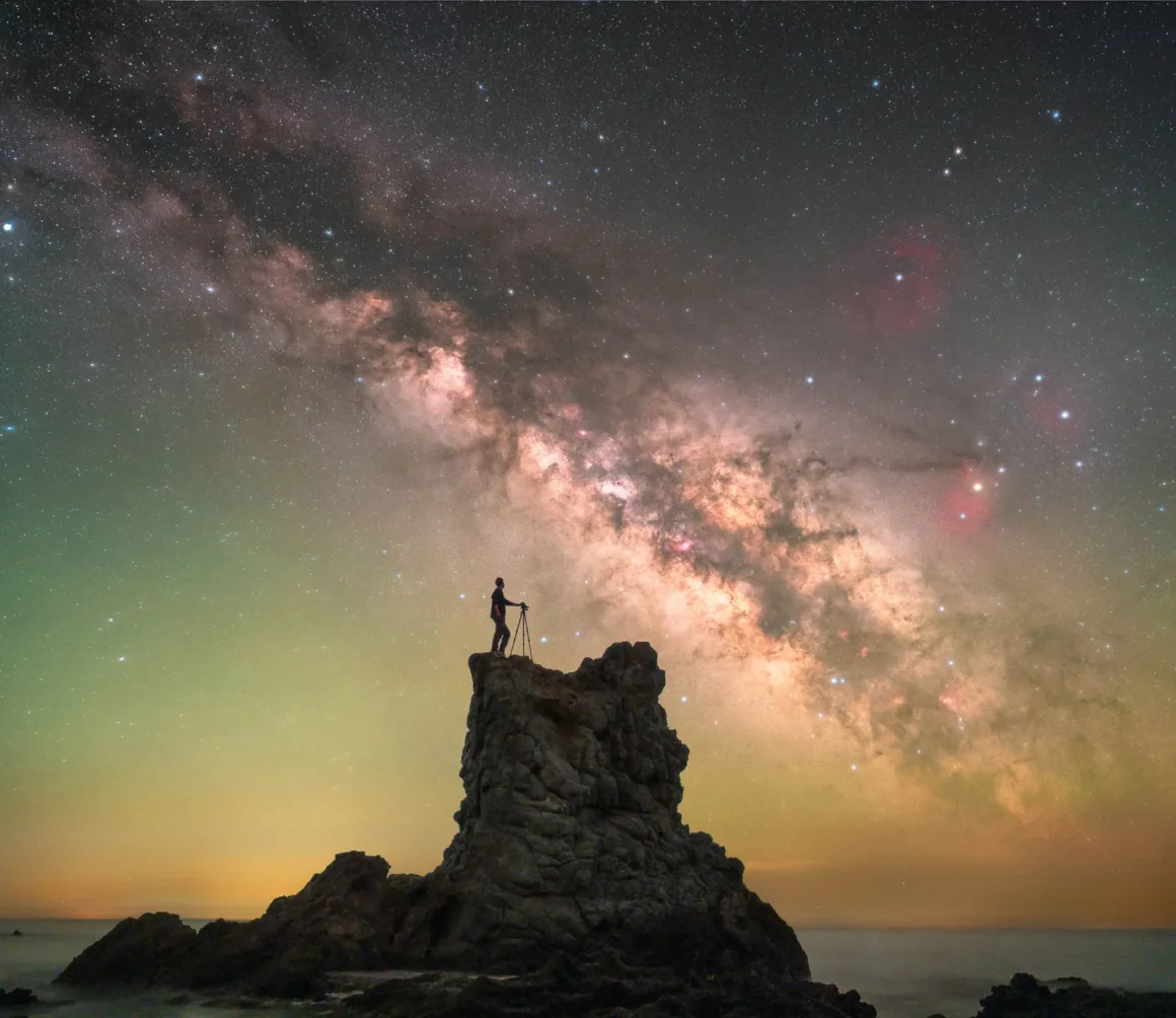 Astronomy Photographer of the Year 2026: Bu Dünyadan Olmayan 31 Kazanan 34 A silhouette of a person standing on a rocky sea stack with a tripod, set against a vibrant Milky Way galaxy and a glowing horizon.