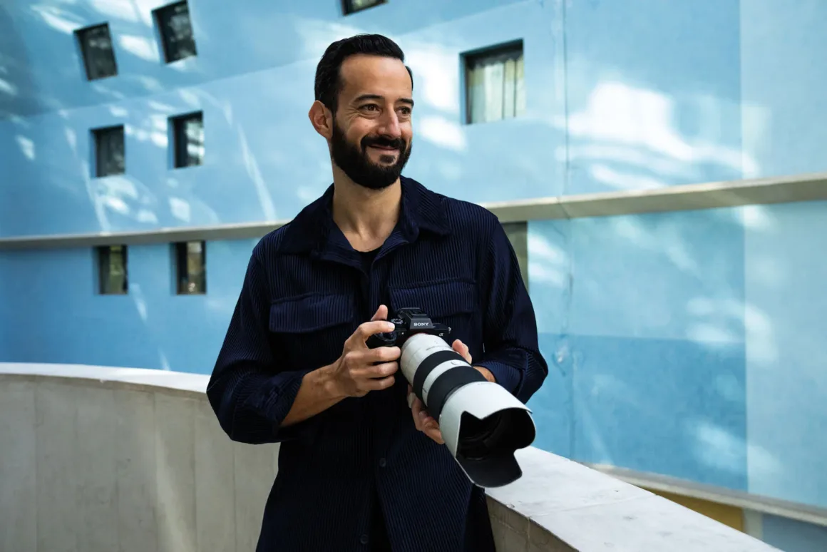 Lacivert düğmeli gömlek giyen sakallı bir adam, kare pencereli mavi bir binanın önünde büyük telefoto objektifli bir fotoğraf makinesi tutuyor.