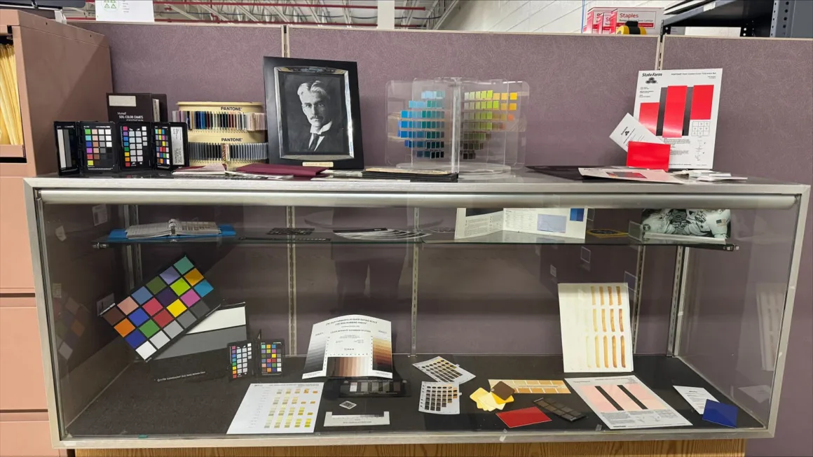 Fotoğrafçıların Renkli Dostu ColorChecker 50 Yaşında 28 Bir vitrinde çeşitli renk kalibrasyon kartları, Pantone örnekleri ve Albert H. Munsell’in çerçeveli portresi bulunuyor.