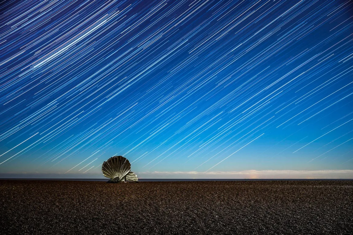 Astronomy Photographer of the Year 2026: Bu Dünyadan Olmayan 31 Kazanan 41 A large metallic shell sculpture stands on a dark pebble beach beneath a night sky filled with long star trails.