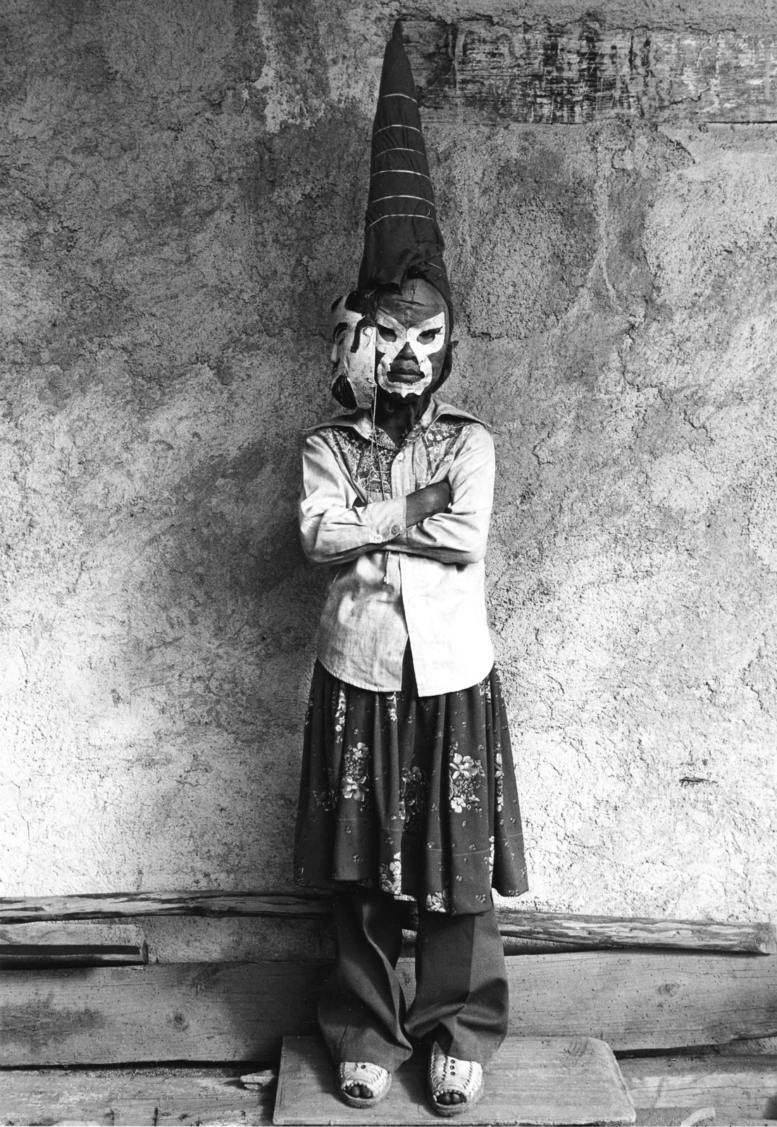 Graciela Iturbide, Leica Hall of Fame’in 15. Üyesi Oldu 20 Bir kişi dokulu bir duvarın önünde kollarını bağlamış şekilde duruyor; uzun konik bir başlık, beyaz işaretli maske, düğmeli gömlek, pantolon üzerine çiçek desenli etek ve sandalet giyiyor.