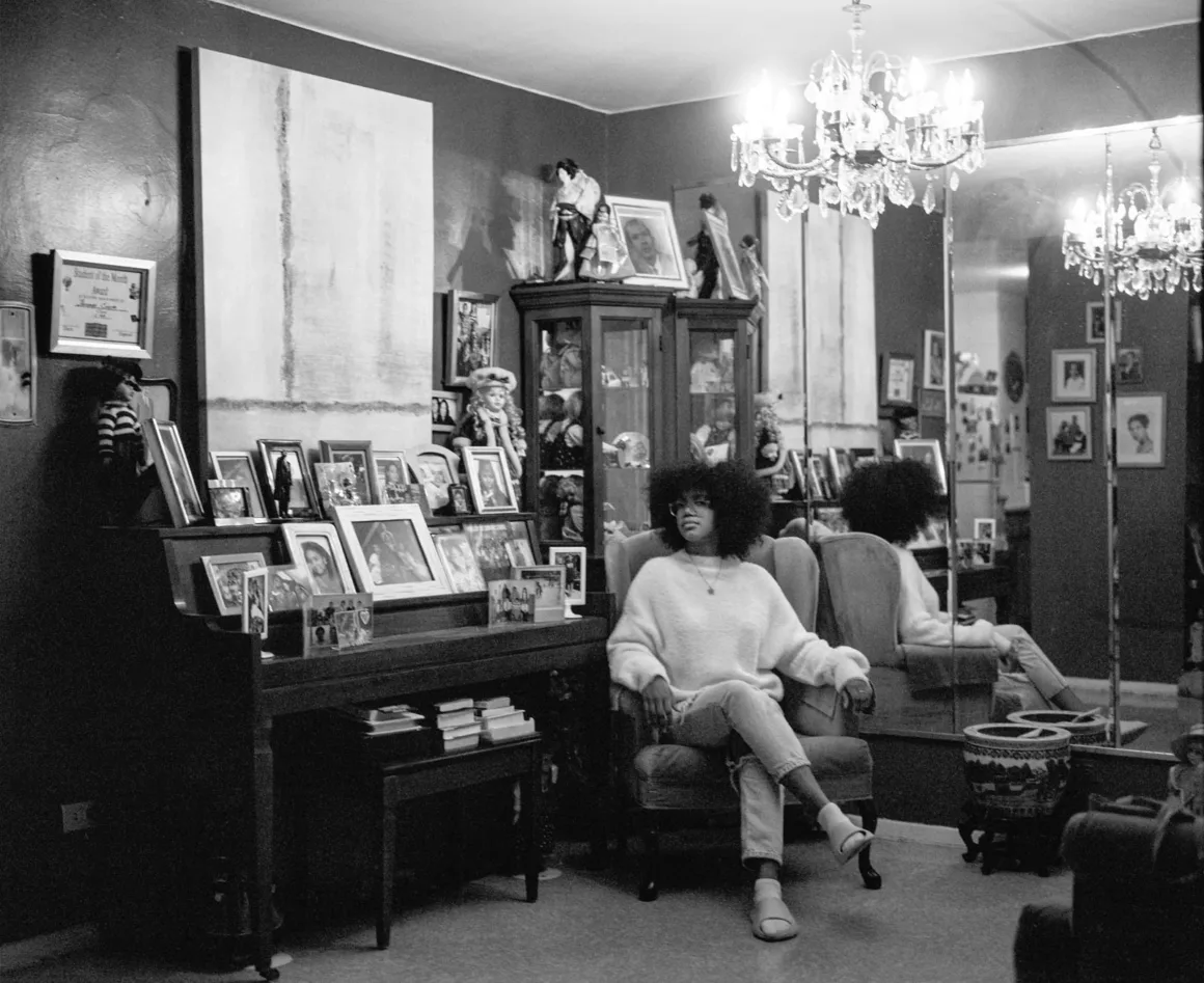 Leica Oskar Barnack Award 2026 Kazananları Açıklandı 28 Afro saçlı bir kişi; çerçeveli fotoğraflar, piyano, cam vitrin ve büyük aynayla dolu bir odada koltukta oturuyor.