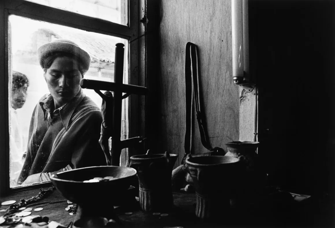 Graciela Iturbide, Leica Hall of Fame’in 15. Üyesi Oldu 23 Şapkalı genç bir adam, bir pencerenin ardından ahşap yüzey üzerindeki seramik kaselere ve haça bakıyor.