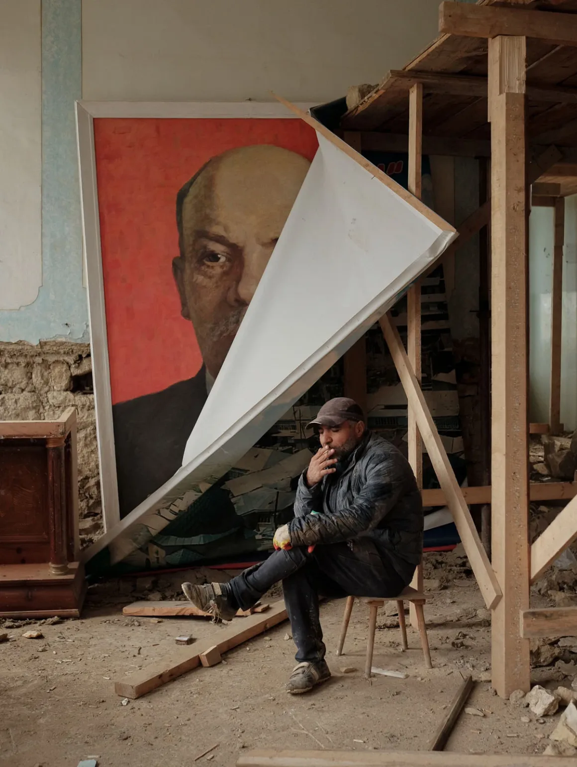 Leica Oskar Barnack Award 2026 Kazananları Açıklandı 22 İş kıyafetleri içindeki bir adam, tozlu ve tamamlanmamış bir odada küçük bir taburede sigara içerken oturuyor; yanında büyük ve kısmen kapatılmış Vladimir Lenin portresi bulunuyor.