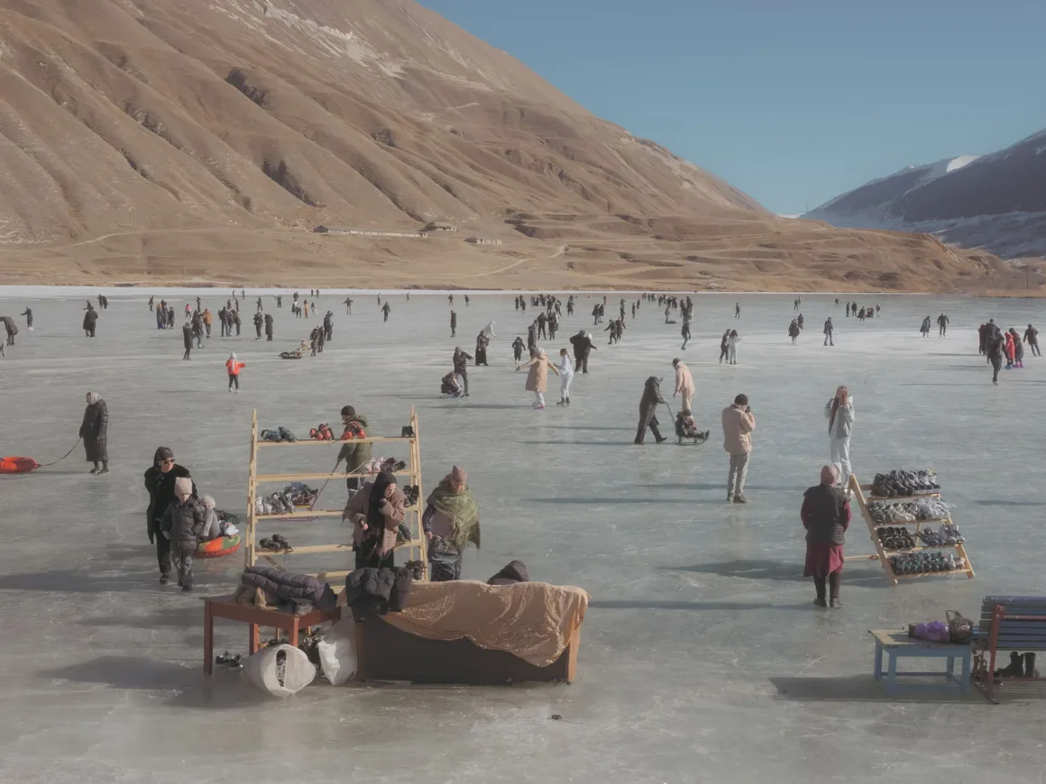 Leica Oskar Barnack Award 2026 Kazananları Açıklandı 24 İnsanlar, çıplak dağlarla çevrili donmuş bir gölde kayıyor ve yürüyor; ahşap raflarda kiralık buz patenleri görülüyor.
