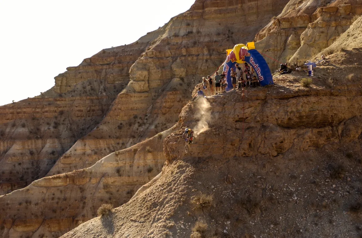 Uçurumun Kenarında Fotoğraf: Red Bull Rampage’in Görünmeyen Yüzü 26 Bir dağ bisikletçisi dik ve kayalık uçurum yüzeyinden aşağı inerken, üst tarafta Red Bull logolu şişme kemerin çevresinde seyirciler toplanmış.