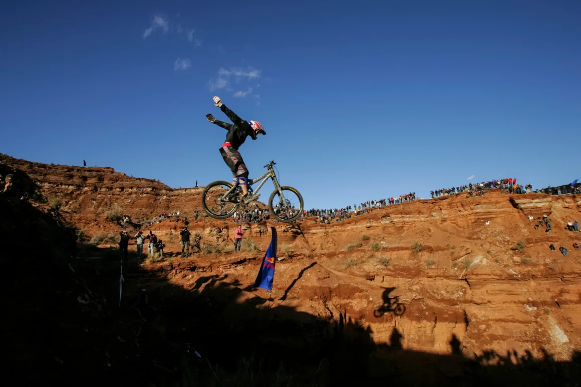 Uçurumun Kenarında Fotoğraf: Red Bull Rampage’in Görünmeyen Yüzü 25 Bir dağ bisikletçisi kayalık kanyonun üzerinde ellerini gidondan bırakarak havada sıçrıyor; sırtta izleyen seyirciler görülüyor.
