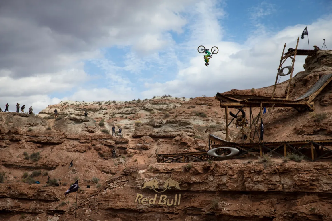 Uçurumun Kenarında Fotoğraf: Red Bull Rampage’in Görünmeyen Yüzü 24 Bir dağ bisikletçisi çöl manzarasındaki ahşap rampadan havalanarak ters takla yapıyor; çevredeki kayalıklardan izleyen seyirciler görülüyor.