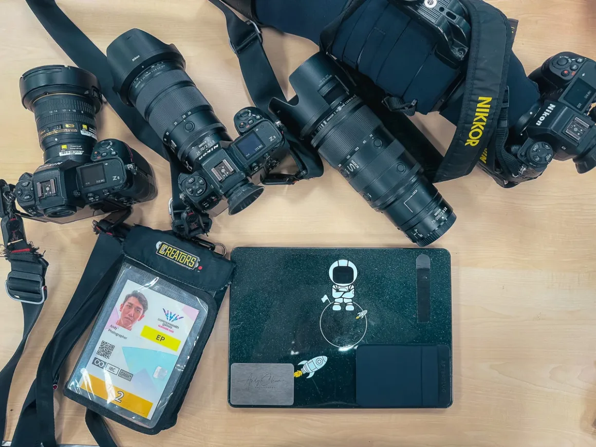 Glasgow 2026’da Bir Spor Fotoğrafçısının Çantasında Neler Vardı? 21 Farklı objektifler takılı üç Nikon kamera, fotoğrafçı akreditasyonu ve üzerinde astronot çıkartması bulunan dizüstü bilgisayar ahşap yüzey üzerinde duruyor.