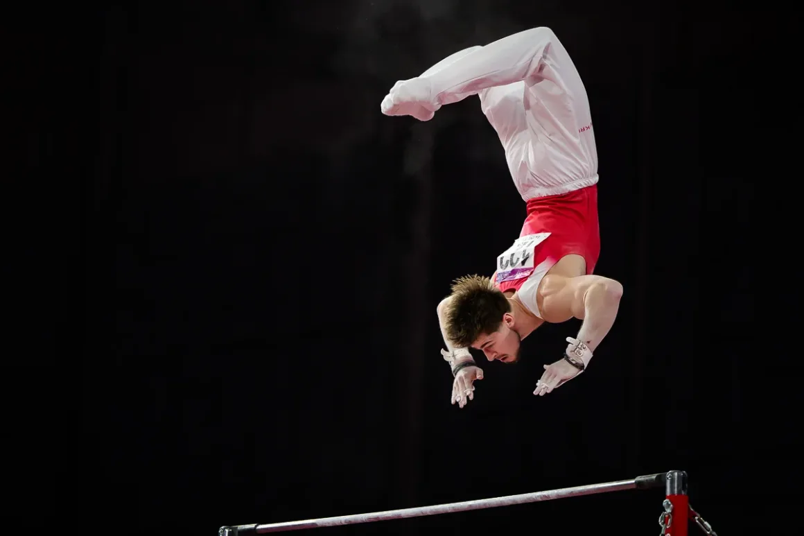Glasgow 2026’da Bir Spor Fotoğrafçısının Çantasında Neler Vardı? 22 Kırmızı-beyaz forma giyen erkek jimnastikçi, koyu arka plan önünde barfikste bırakma hareketi yapıyor.