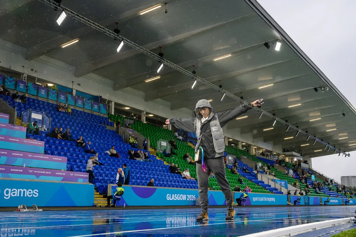 Glasgow 2026’da Bir Spor Fotoğrafçısının Çantasında Neler Vardı? 28 Kapüşonlu yağmurluk giyen kişi, mavi ve yeşil tribünlerin bulunduğu stadyumda ıslak mavi koşu pistinde kollarını açmış halde duruyor.