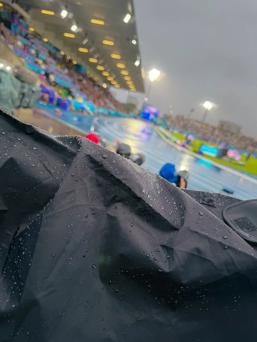 Glasgow 2026’da Bir Spor Fotoğrafçısının Çantasında Neler Vardı? 29 Ön plandaki siyah su geçirmez kumaş üzerinde yağmur damlaları, arka planda ise stadyum ışıkları altında bulanık pist ve seyirciler görülüyor.