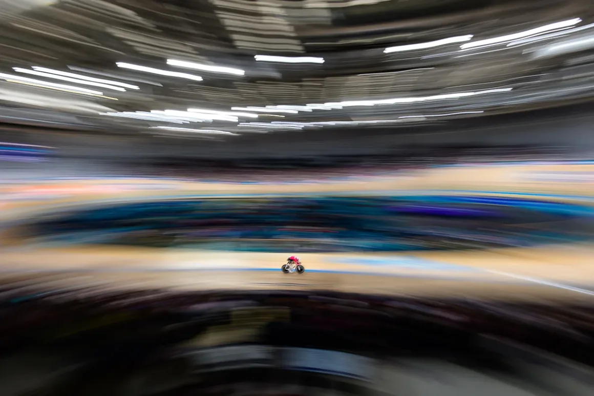 Glasgow 2026’da Bir Spor Fotoğrafçısının Çantasında Neler Vardı? 25 Kırmızı forma giyen bisikletçi, yoğun hareket bulanıklığıyla çevrili velodrom pistinde yarışıyor.