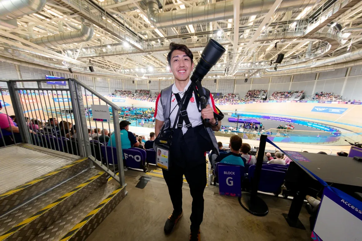 Glasgow 2026’da Bir Spor Fotoğrafçısının Çantasında Neler Vardı? 34 Gülümseyen fotoğrafçı stadyumun medya bölümünde, omzunda büyük telefoto objektif ve üzerinde kamera taşıma sistemiyle duruyor.
