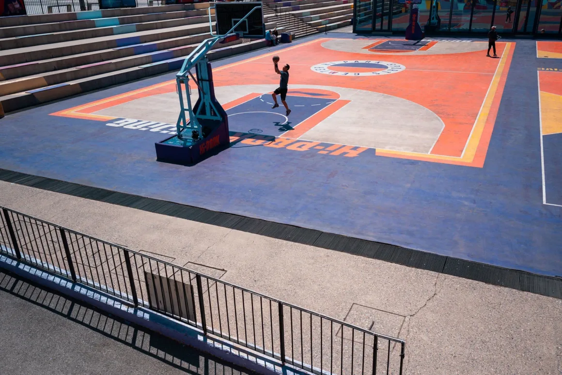 TTArtisan AF 24mm f/3.8 Mini: Sony E Mount için 57 Gramlık Pancake Objektif 24 A person jumps to shoot a basketball at an outdoor court with orange and blue painted surfaces and tiered concrete seating.