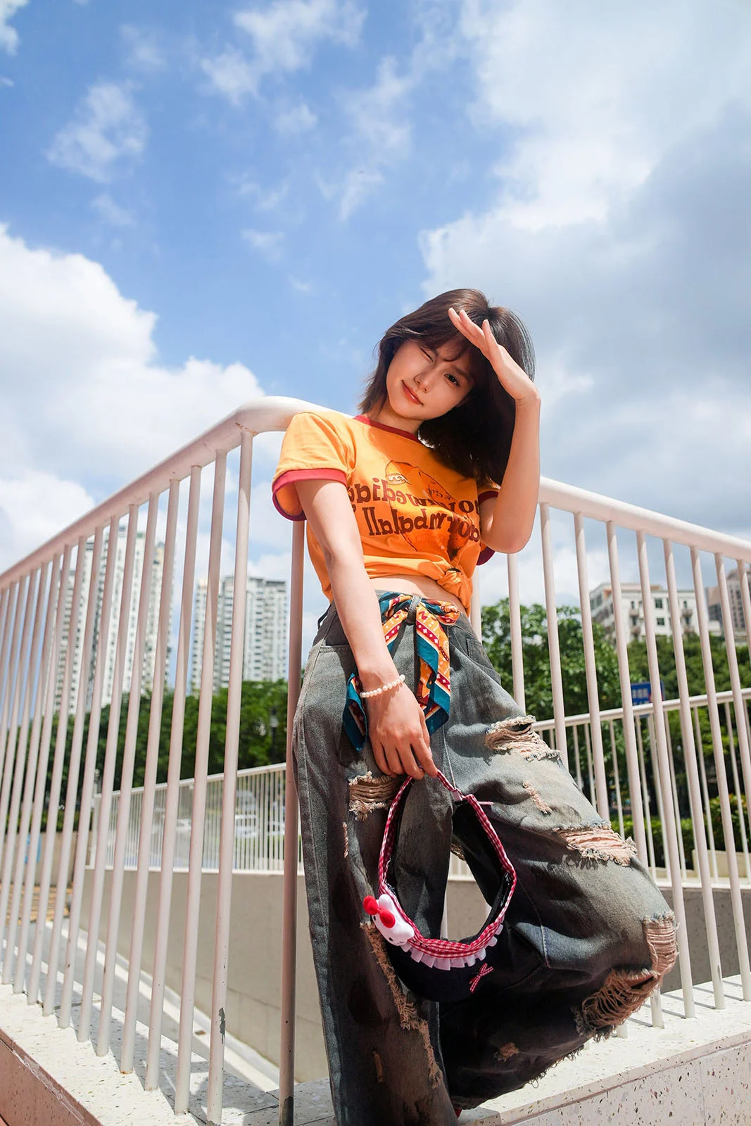 TTArtisan AF 24mm f/3.8 Mini: Sony E Mount için 57 Gramlık Pancake Objektif 25 A young woman with short dark hair poses outdoors against a white railing, wearing an orange t-shirt, distressed jeans, and holding a small patterned bag.