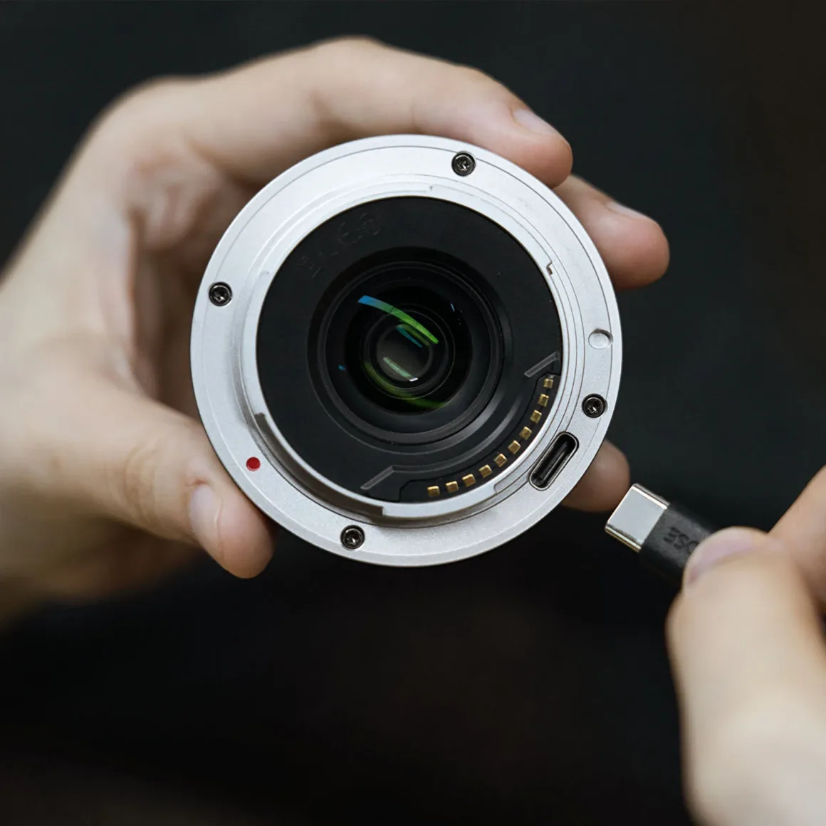 TTArtisan AF 24mm f/3.8 Mini: Sony E Mount için 57 Gramlık Pancake Objektif 22 Bir kişi objektifi gümüş renkli bayonetinden tutarken diğer eliyle objektifin yan tarafındaki bağlantı noktasına USB-C kablosu takmaya hazırlanıyor.