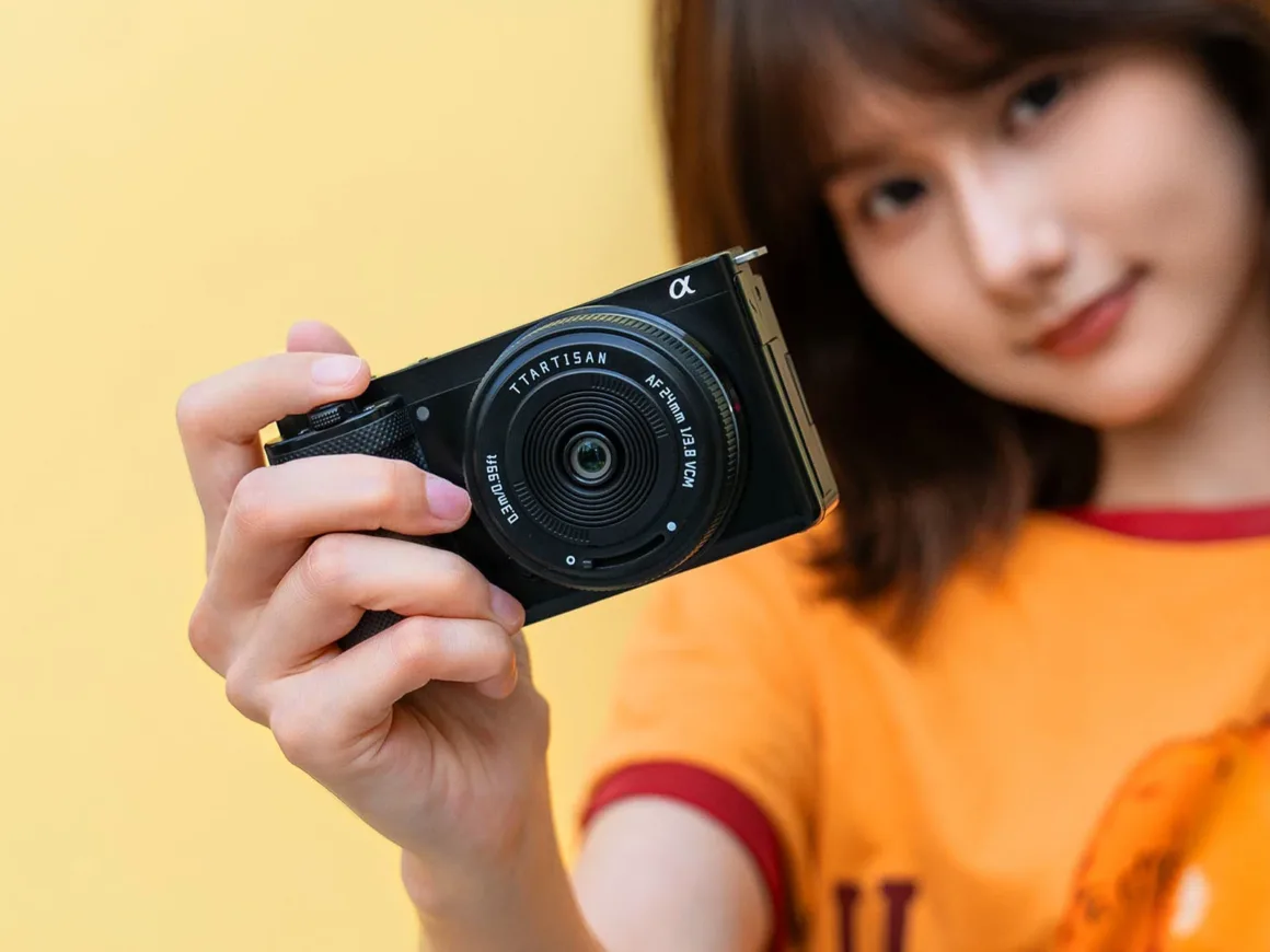TTArtisan AF 24mm f/3.8 Mini: Sony E Mount için 57 Gramlık Pancake Objektif 20 Omuz hizasında kahverengi saçları olan bir kişi, sarı arka plan önünde TTArtisan AF 24mm f/1.8 VCM objektif takılı siyah dijital fotoğraf makinesini kameraya doğru tutuyor.
