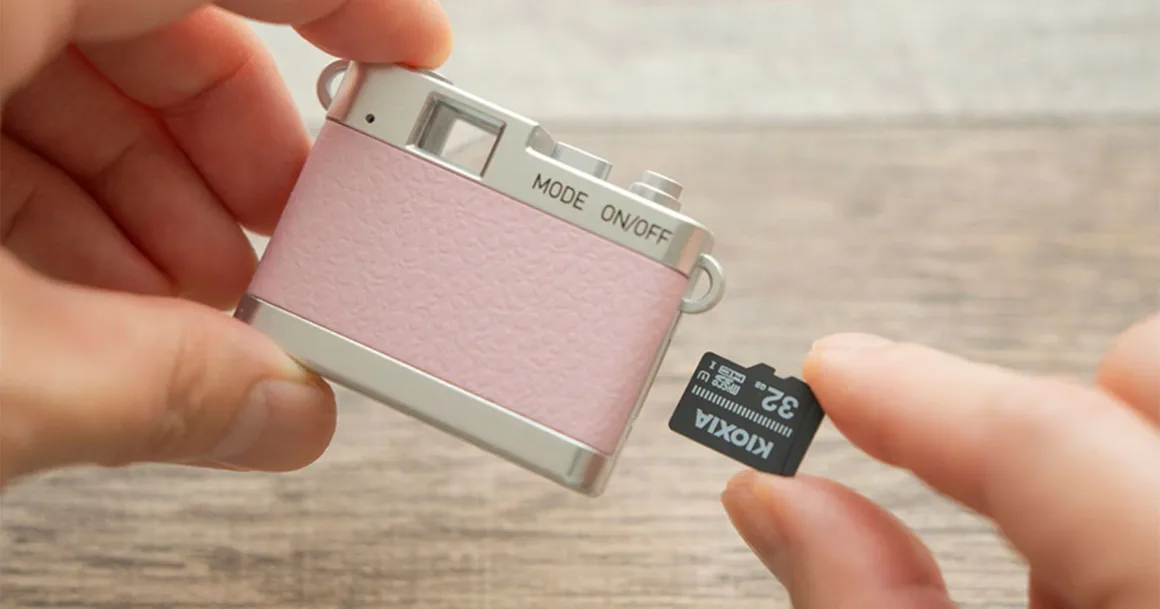 Kenko Tokina Pieni III: Kolye Gibi Taşınabilen 18 Gramlık Mini Kamera 23 Bir kişi bir elinde küçük, pembe ve gümüş renkli oyuncak görünümlü kamera, diğer elinde ise Kioxia 32GB microSD kart tutuyor.