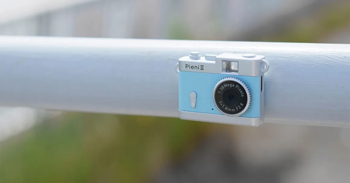 Kenko Tokina Pieni III: Kolye Gibi Taşınabilen 18 Gramlık Mini Kamera 21 Açık mavi renkli, oyuncak benzeri küçük Pieni III dijital fotoğraf makinesi beyaz bir korkuluğun üzerinde duruyor.