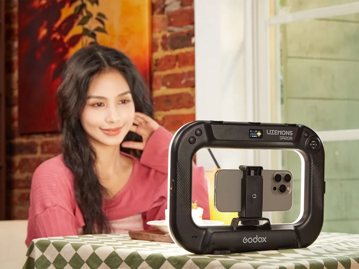 Godox Litemons SR20R, Telefon ve Gimbal Kameralar için RGB Işık Sunuyor 20 Pembe gömlek giyen bir kadın, ortasında akıllı telefon bulunan Godox Litemons SR20R ışığın arkasında masada oturuyor.