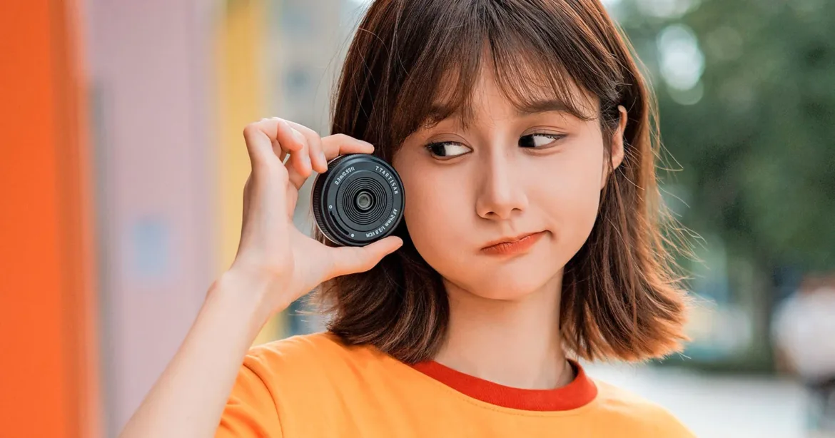 A young woman with shoulder-length brown hair and an orange shirt holds a small, black camera lens up to her cheek while looking to the side.