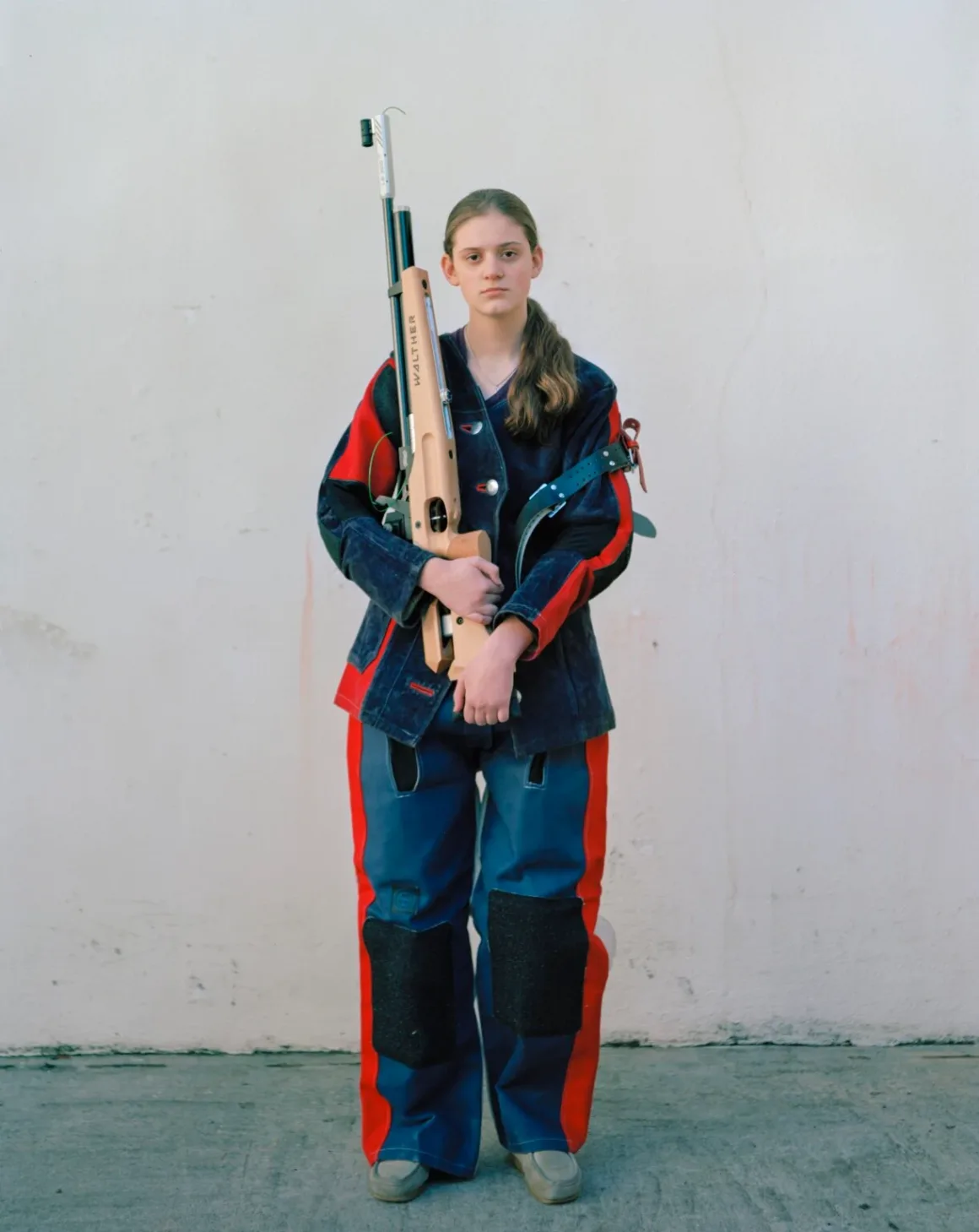 Eva Verbeeck, Amerika’nın Genç Kızlarını 6 Yıl Boyunca Fotoğrafladı 17 Mavi ve kırmızı atıcılık kıyafeti giyen genç bir kişi, sade bir duvarın önünde Walther havalı tüfeğini dikey olarak tutuyor.